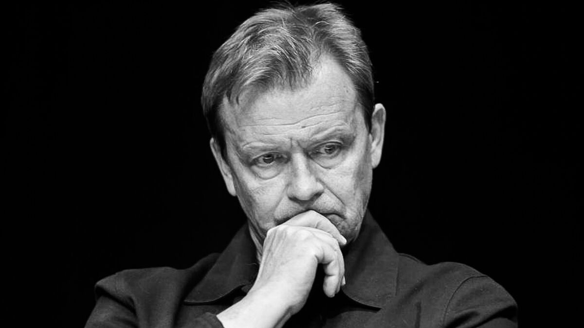 Jan Frycz jest pokazany na czarno-białym portrecie, z głową lekko pochyloną i wzrokiem skierowanym w dół. Aktor opiera zaciśniętą dłoń o usta i ma zamyślony, poważny wyraz twarzy. W tle widoczna jest jednolita, czarna przestrzeń kontrastująca z oświetloną twarzą i dłonią.