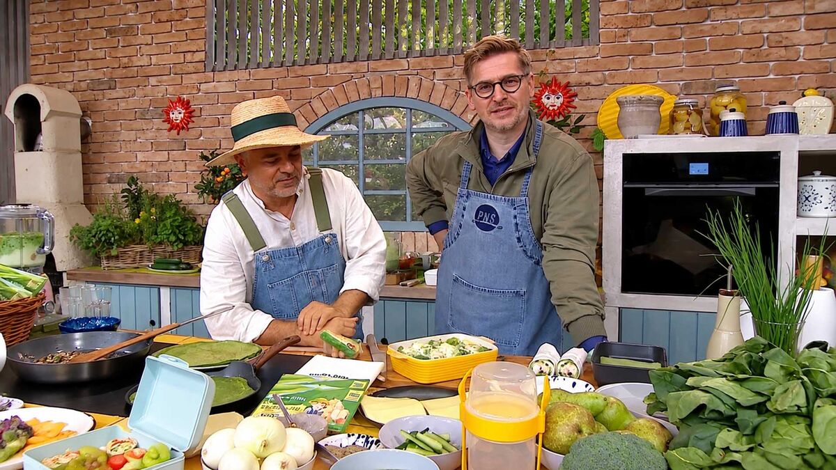 Po prawej Tomasz Królikowski stoi przy kuchennym blacie obok mężczyzny w słomkowym kapeluszu, a obaj mają na sobie niebieskie fartuchy. Przed nimi ustawiono przygotowane potrawy, patelnie, pojemniki oraz świeże warzywa, w tym szpinak, brokuły, cebulę i ogórki. W tle znajdują się ceglana ściana, łukowe okno, rośliny, piekarnik oraz naczynia ustawione na półkach.