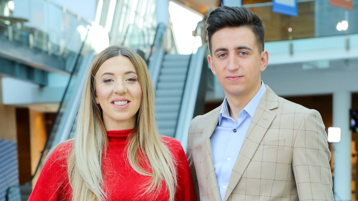 Małgorzata i Paweł Borysewicz stoją obok siebie w nowoczesnym wnętrzu z ruchomymi schodami w tle. Oboje patrzą w stronę aparatu, prezentując się w eleganckich, schludnych stylizacjach. Jasne, przestronne otoczenie nadaje scenie profesjonalny i świeży charakter.