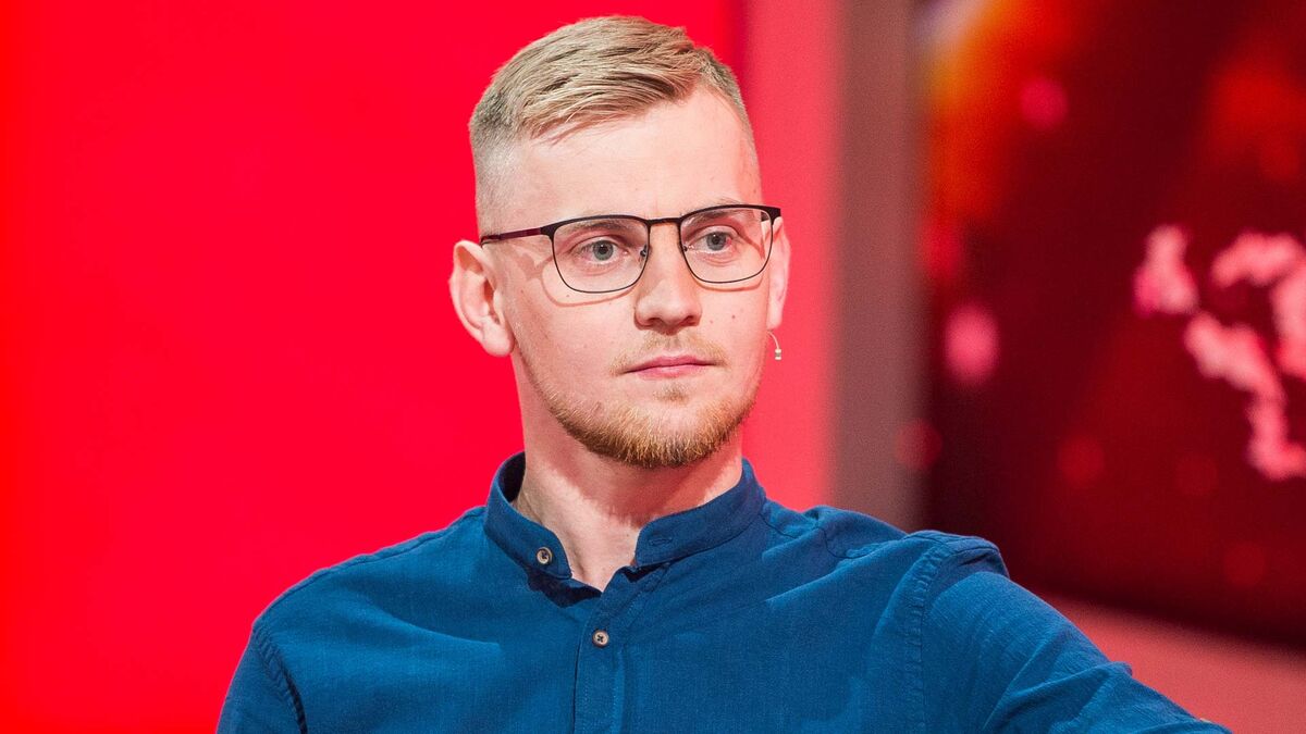 Karol z programu „Rolnik szuka żony” siedzi w studiu, ubrany w niebieską koszulę i czarne okulary. Mężczyzna jest pokazany w zbliżeniu, a przy jego twarzy widoczny jest niewielki mikrofon. W tle znajdują się czerwone elementy scenografii i rozmyte światła.