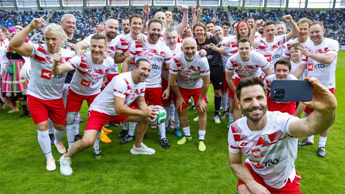 Grupa zawodników w biało-czerwonych strojach stoi na murawie stadionu i wykonuje wspólne zdjęcie typu selfie, trzymane przez jednego z uczestników. W tle widoczne są trybuny z publicznością oraz elementy infrastruktury stadionowej.