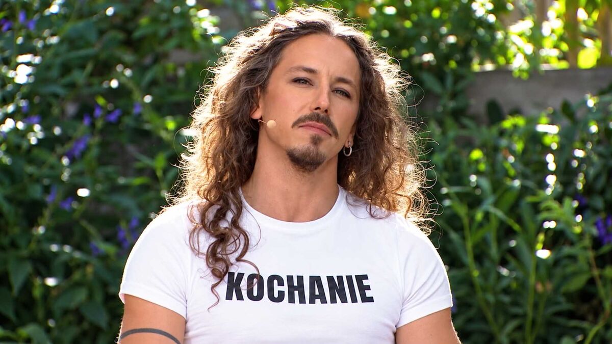 Michał Szpak ma długie falowane włosy, biały T-shirt z napisem „KOCHANIE” oraz mikrofon przy policzku. Na ramieniu widoczny jest czarny pasek, a w uchu niewielkie kółko. Tło tworzą zielone liście i fioletowe kwiaty oświetlone światłem słonecznym.
