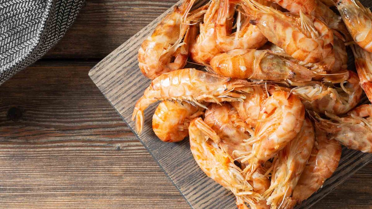 Na drewnianej desce leży porcja krewetek w pancerzach, ułożonych w stos na ciemnym blacie. Kadr pokazuje zbliżenie na pomarańczowo-różowe skorupki i czułki, a w lewym górnym rogu widać fragment metalowego naczynia. Tło stanowi drewniana powierzchnia z wyraźnym usłojeniem.