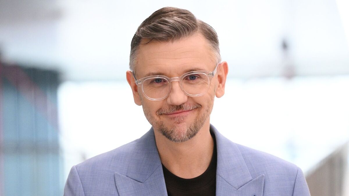 Krzysztof Czeczot patrzy wprost w obiektyw i lekko się uśmiecha. Ma na sobie jasną marynarkę, czarną koszulkę oraz przezroczyste okulary w cienkich oprawkach. W tle widać jasną, nowoczesną przestrzeń z rozmytymi elementami architektury.