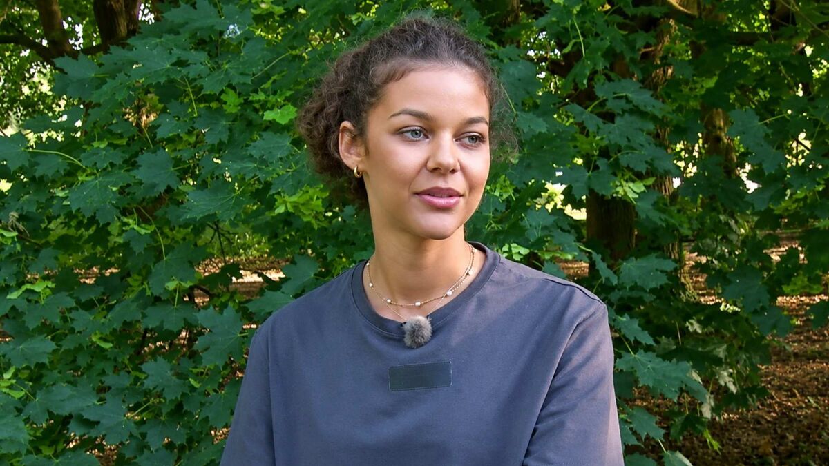 Emilia Dankwa siedzi w plenerze na tle gęstych zielonych drzew. Ma na sobie szarą koszulkę i przypięty do niej mikrofon, a głowę zwraca lekko w bok. Zdjęcie wykonano w ciągu dnia, w otoczeniu parku lub zadrzewionego terenu.