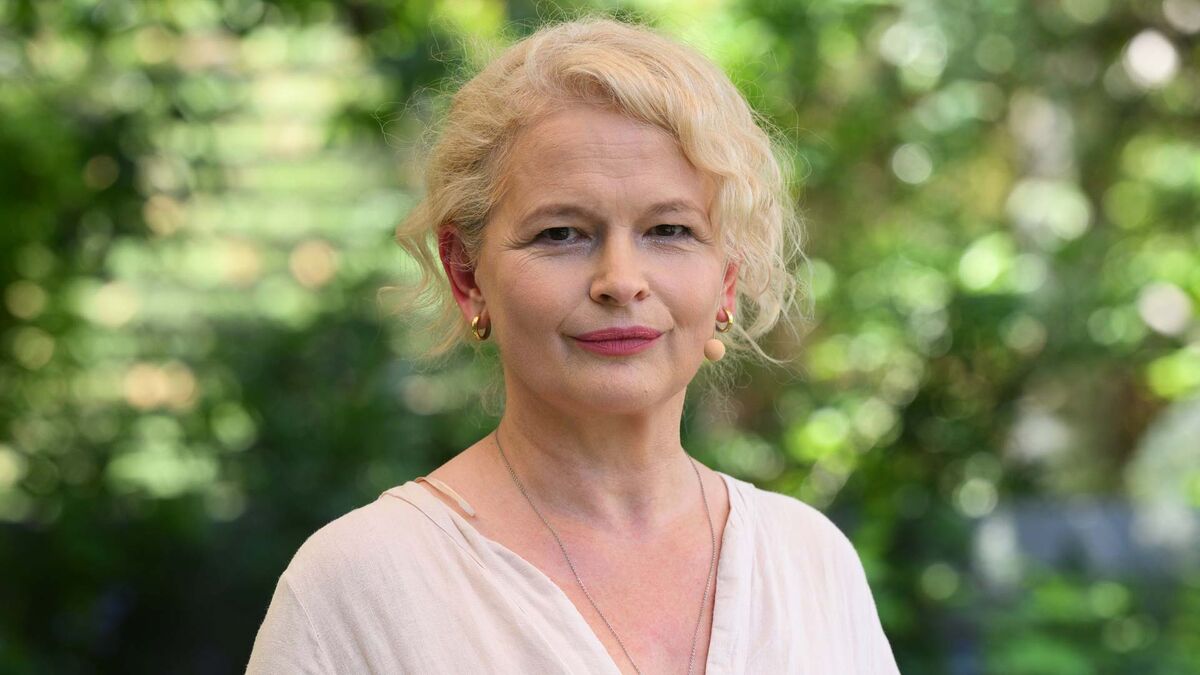 Na zdjęciu widoczna jest Dominika Ostałowska w ujęciu portretowym. Ma krótkie blond włosy, jasną bluzkę oraz niewielkie kolczyki i naszyjnik. W tle znajduje się rozmyta zieleń ogrodu.