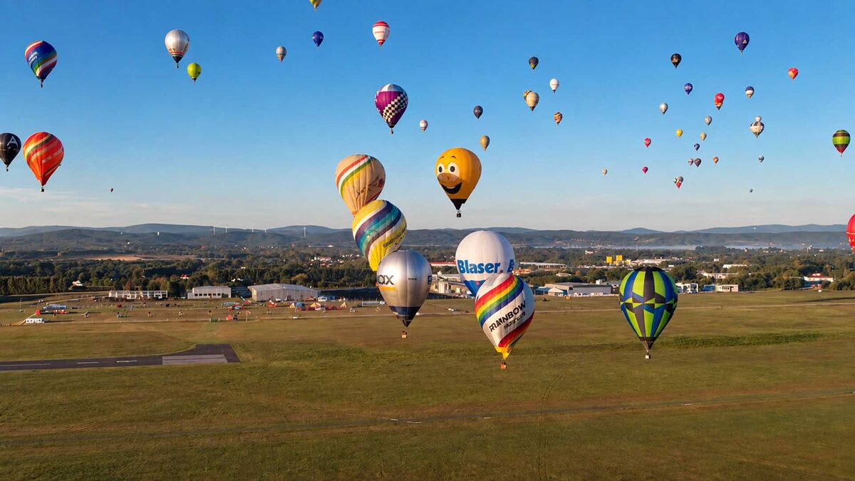 Kilkadziesiąt balonów unosi się nad terenem zawodów balonowych w Krośnie. Balony mają różne kolory, wzory i kształty, w tym jeden z dużą rysunkową twarzą. Poniżej rozciąga się trawiasty teren z zabudowaniami, a w oddali widoczne są wzgórza.